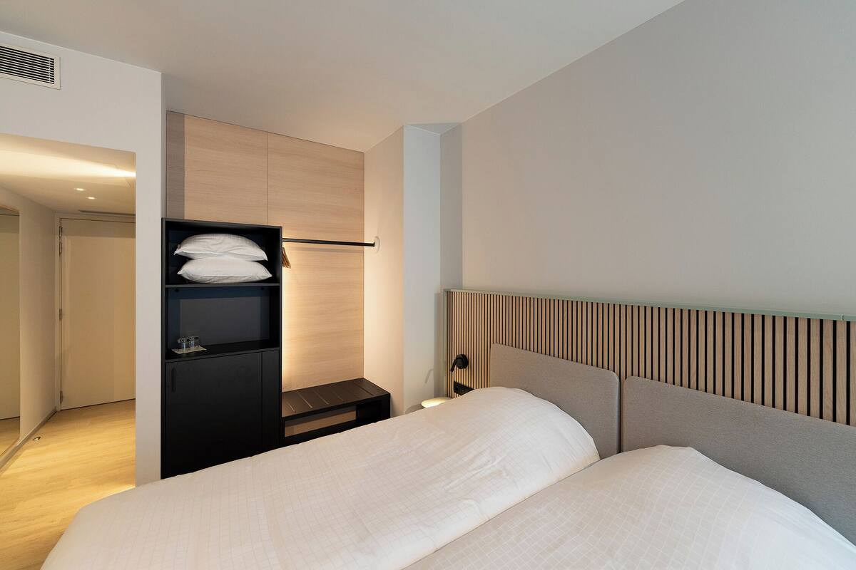 A modern hotel room with a large bed, a bedside table, a wardrobe, and a headboard with vertical wooden slats.