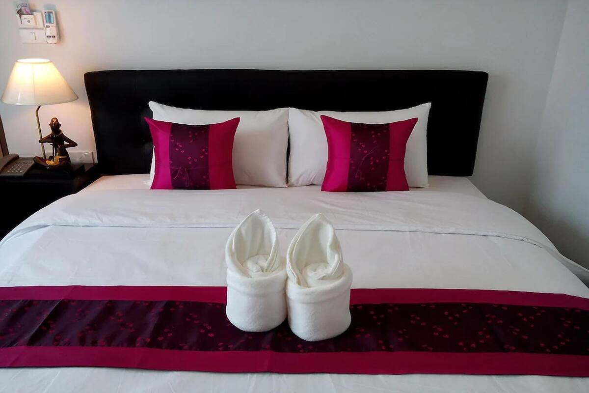 Uma cama bem arrumada com almofadas brancas e rosa, um espalha cama vermelho e preto, e um par de toalhas brancas colocadas sobre a cama.