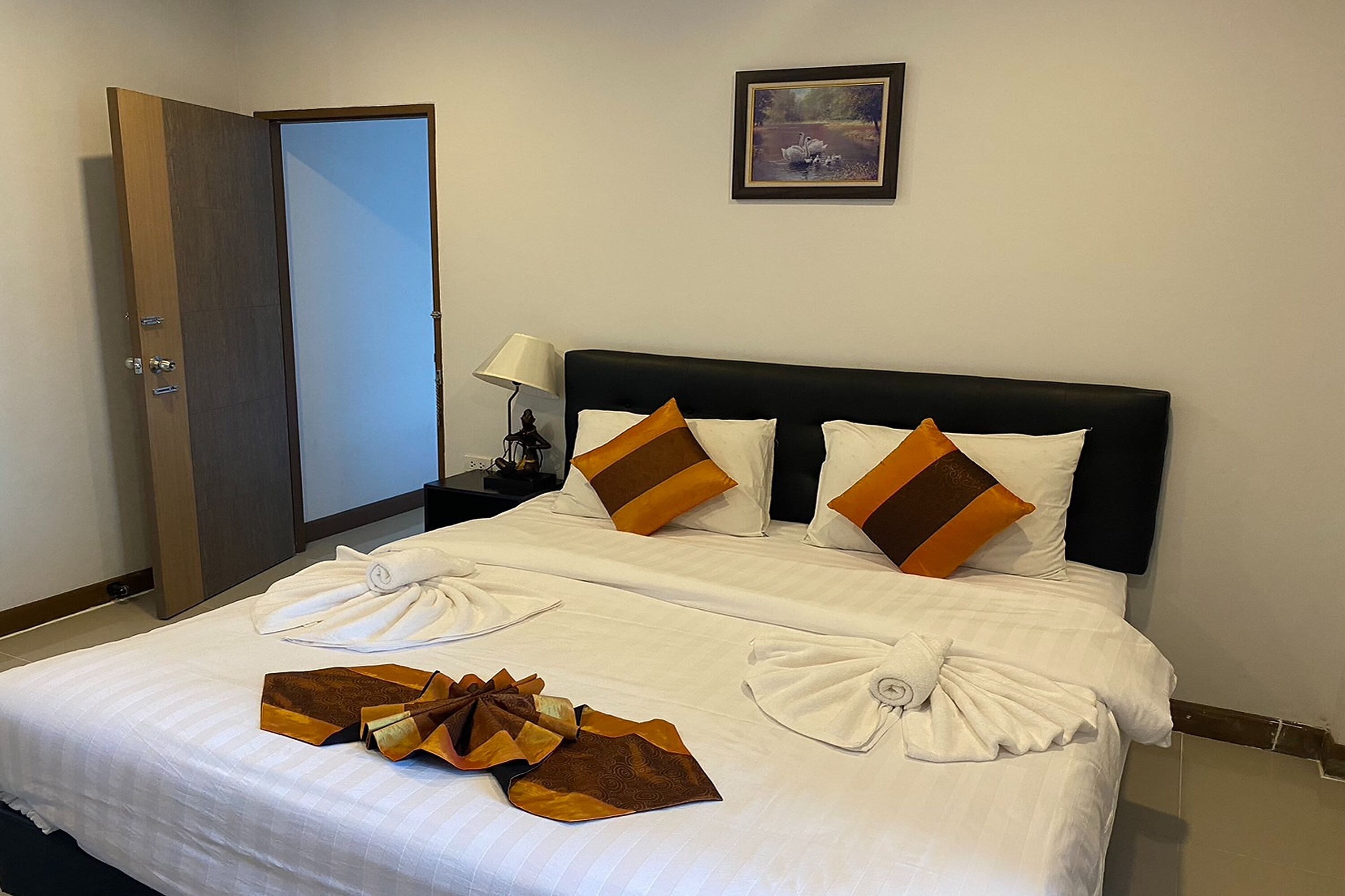 Cama bem arrumada com lençóis brancos e almofadas decorativas castanhas, um quadro na parede e uma porta à esquerda.