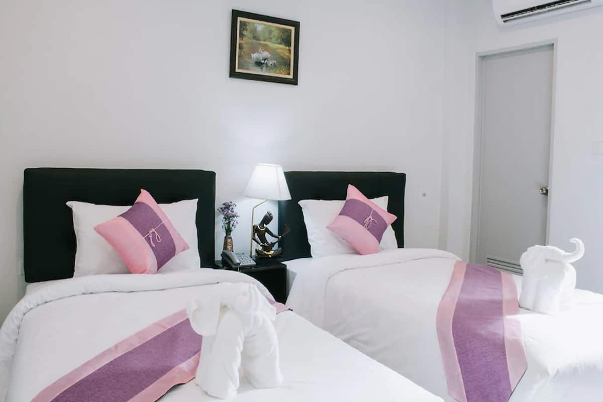 Quarto de hotel com duas camas, cada uma com roupa de cama branca e detalhes em rosa. Há uma mesa de cabeceira com uma luminária e uma pequena estátua. Um quadro emoldurado está pendurado na parede acima das camas.