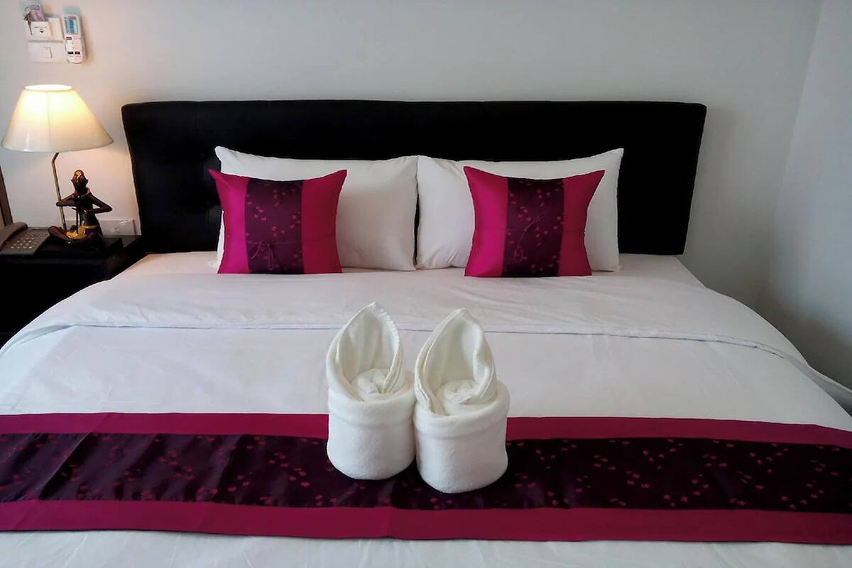Cama bem arrumada com almofadas brancas e cor-de-rosa, cabeceira escura e um par de toalhas brancas sobre um cobreleito vermelho escuro.