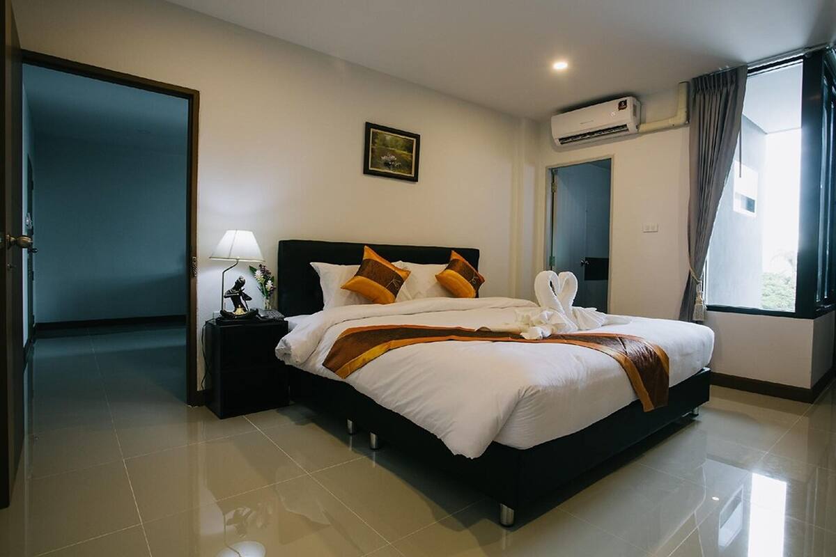 Quarto de hotel com uma cama grande, uma mesa de cabeceira com abajur, ar condicionado na parede e uma janela com cortinas.