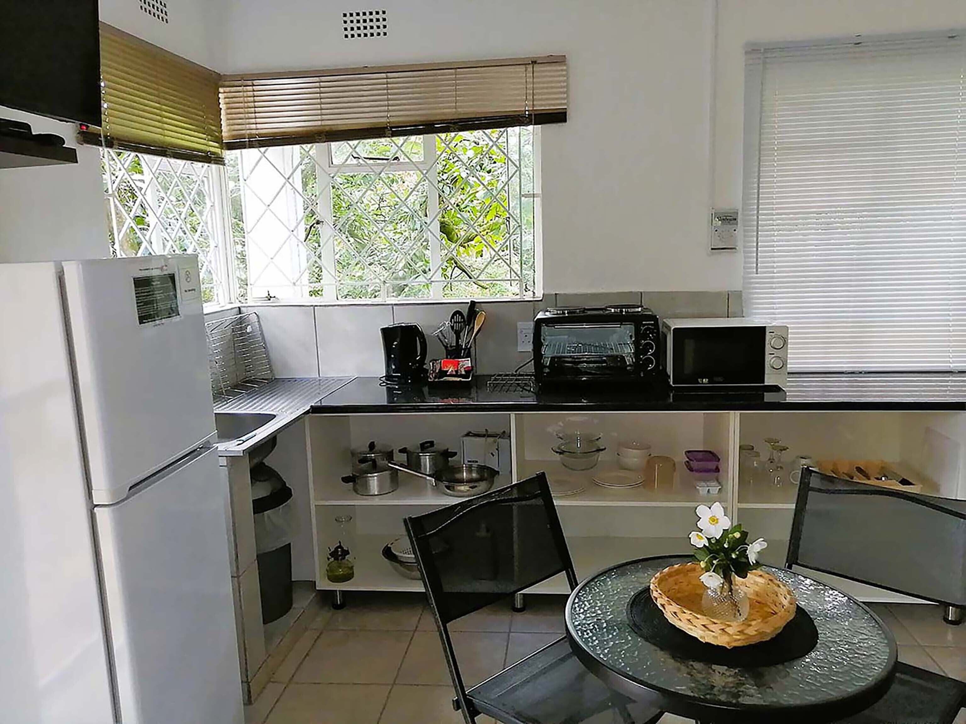 A compact kitchen with a refrigerator, a toaster, a microwave, and a sink. There is a window with blinds, a small dining table with chairs, and a decorative flower arrangement.