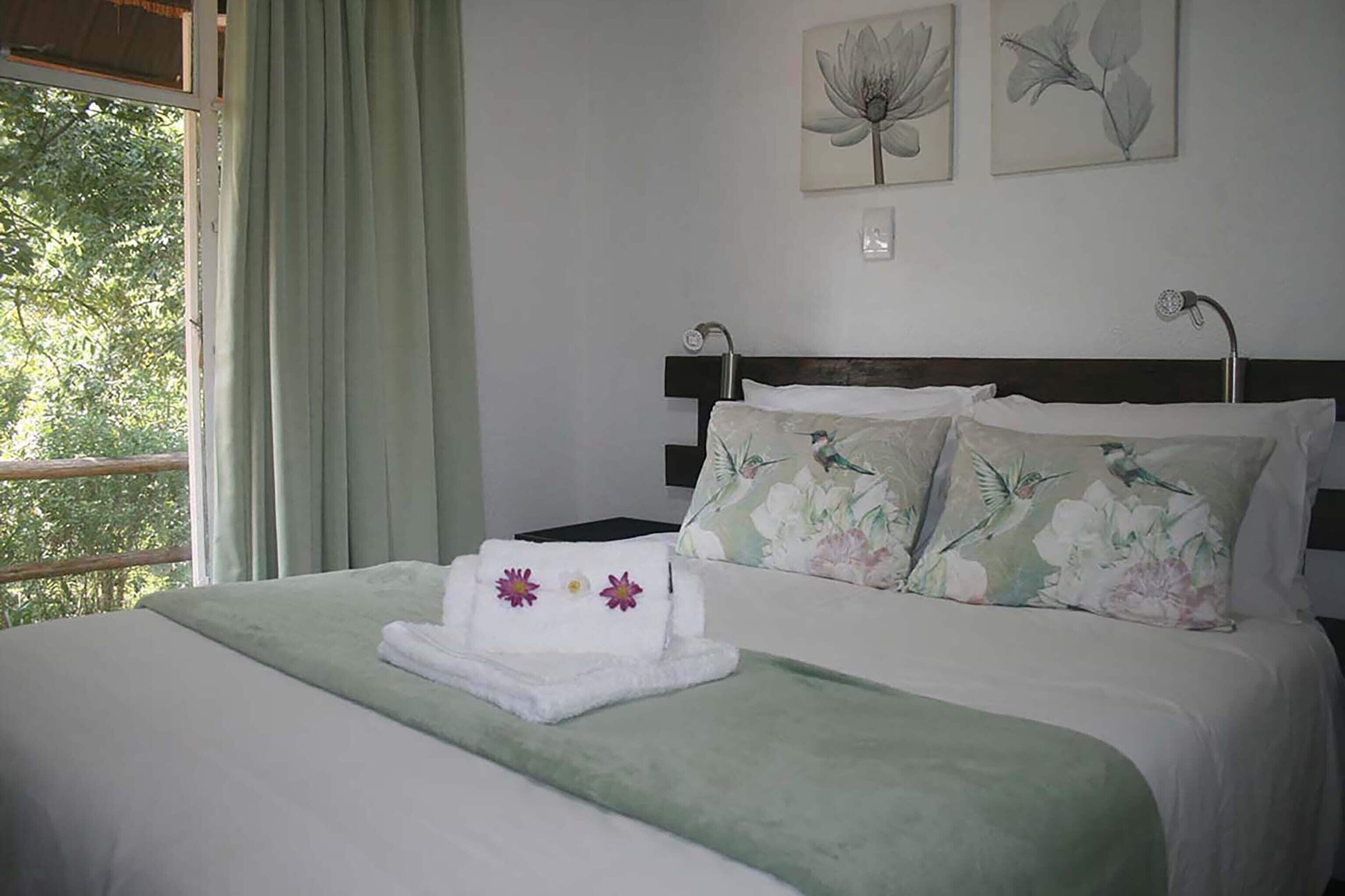 Ein ordentlich bezogenes Bett mit floralen Kissen und einer grünen Tagesdecke, ein Fenster mit grünen Vorhängen und zwei Blumenbilder an der Wand.