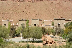 A desert landscape with traditional architecture, including multiple buildings with arched windows and flat roofs, surrounded by sparse vegetation. - Kasbah Hotel Camping Jurassique (Lkheng)