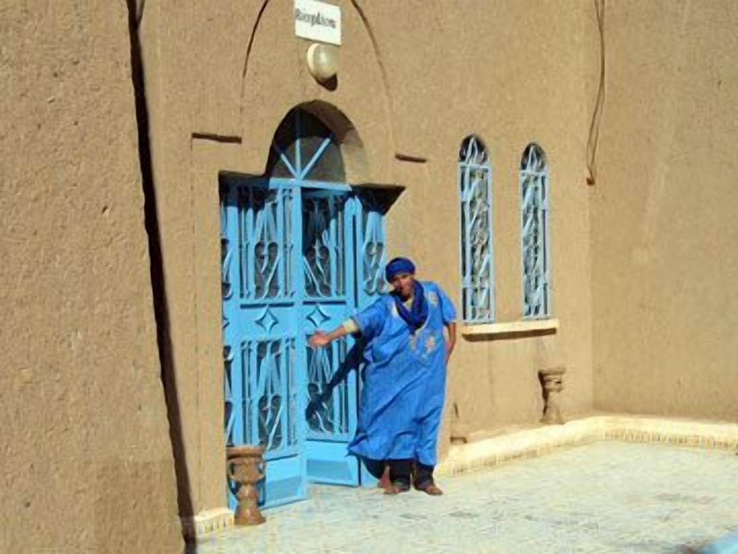 A person in blue attire standing in front of a building with a blue door and arched windows.