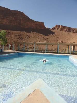A swimming pool with clear blue water, surrounded by a wooden deck and a rocky cliff in the background. - Kasbah Hotel Camping Jurassique (Lkheng)