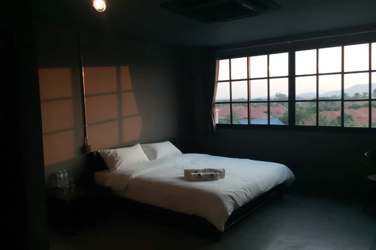 A dimly lit hotel room with a large window, a bed, and a nightstand with a bottle.