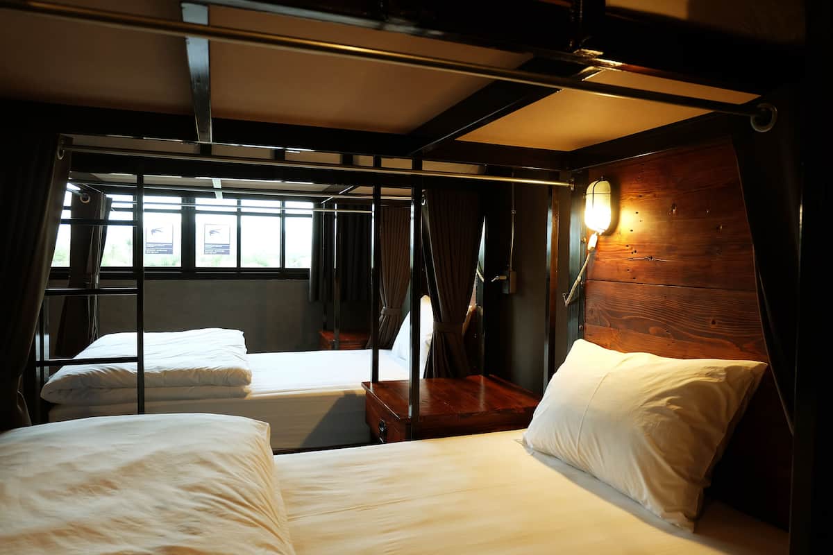 A cabin with two beds, a wooden headboard, a window with curtains, and a wall-mounted light.