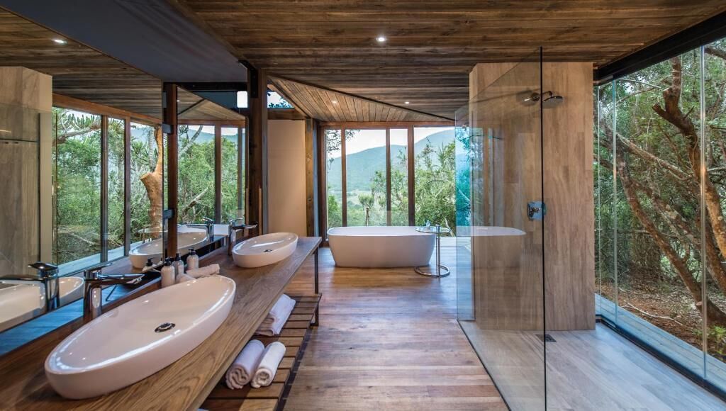 A modern bathroom with a large sink, a bathtub, and a shower area, with large windows offering a view of the outdoors.