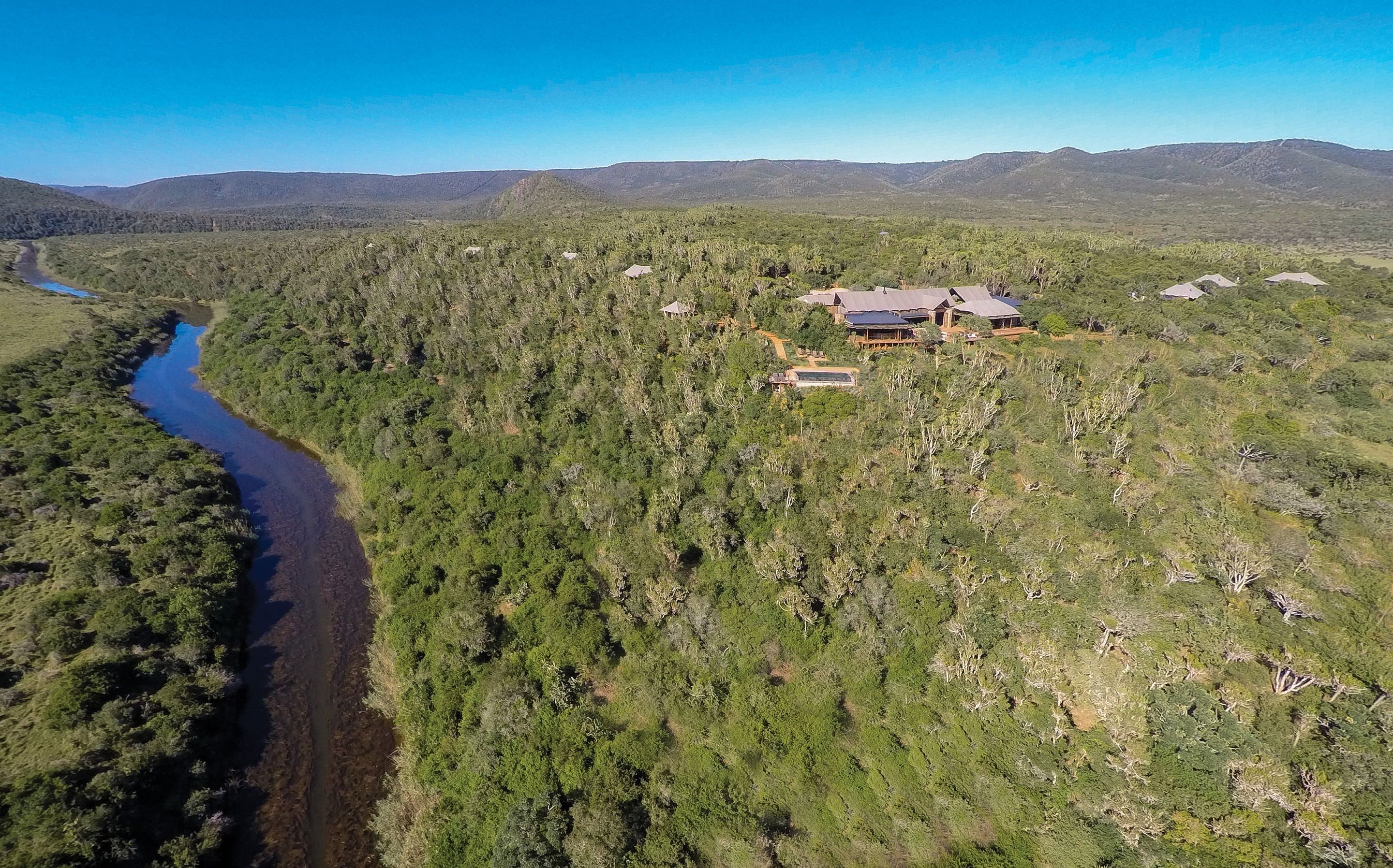 An aerial view of a remote, forested area with a river, a small cluster of buildings with a large house, and distant mountains.