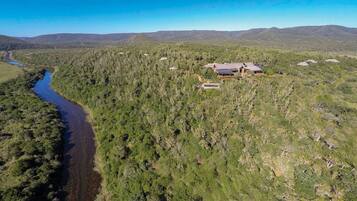 An aerial view of a remote, forested area with a river, a small cluster of buildings with a large house, and distant mountains.