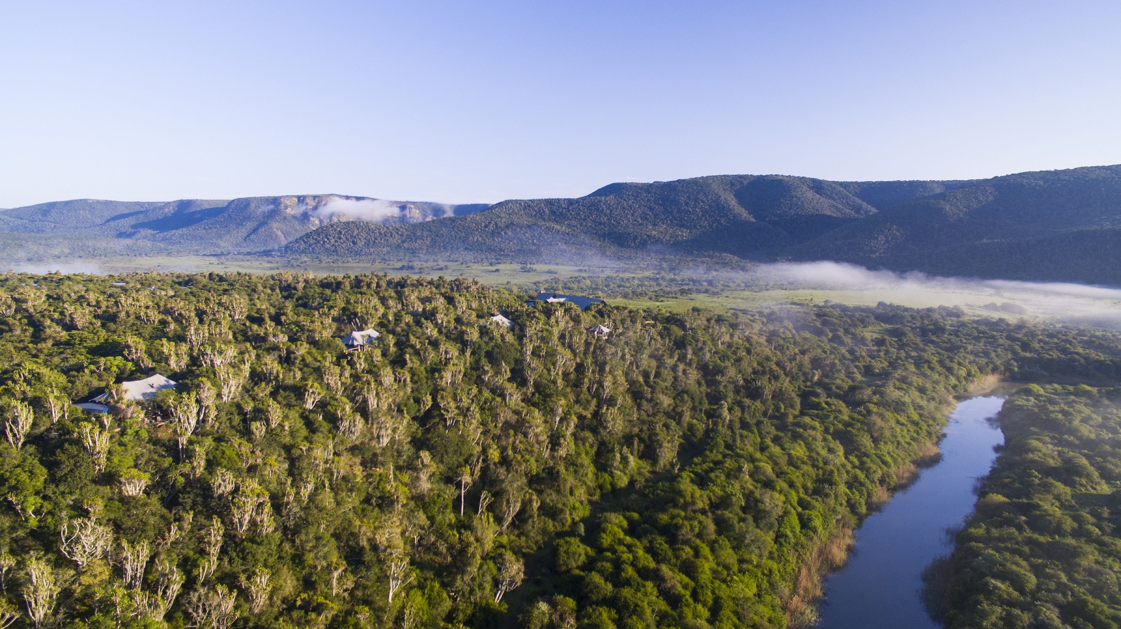An aerial view of a forested area with a river, mountains in the background, and several small buildings scattered throughout.