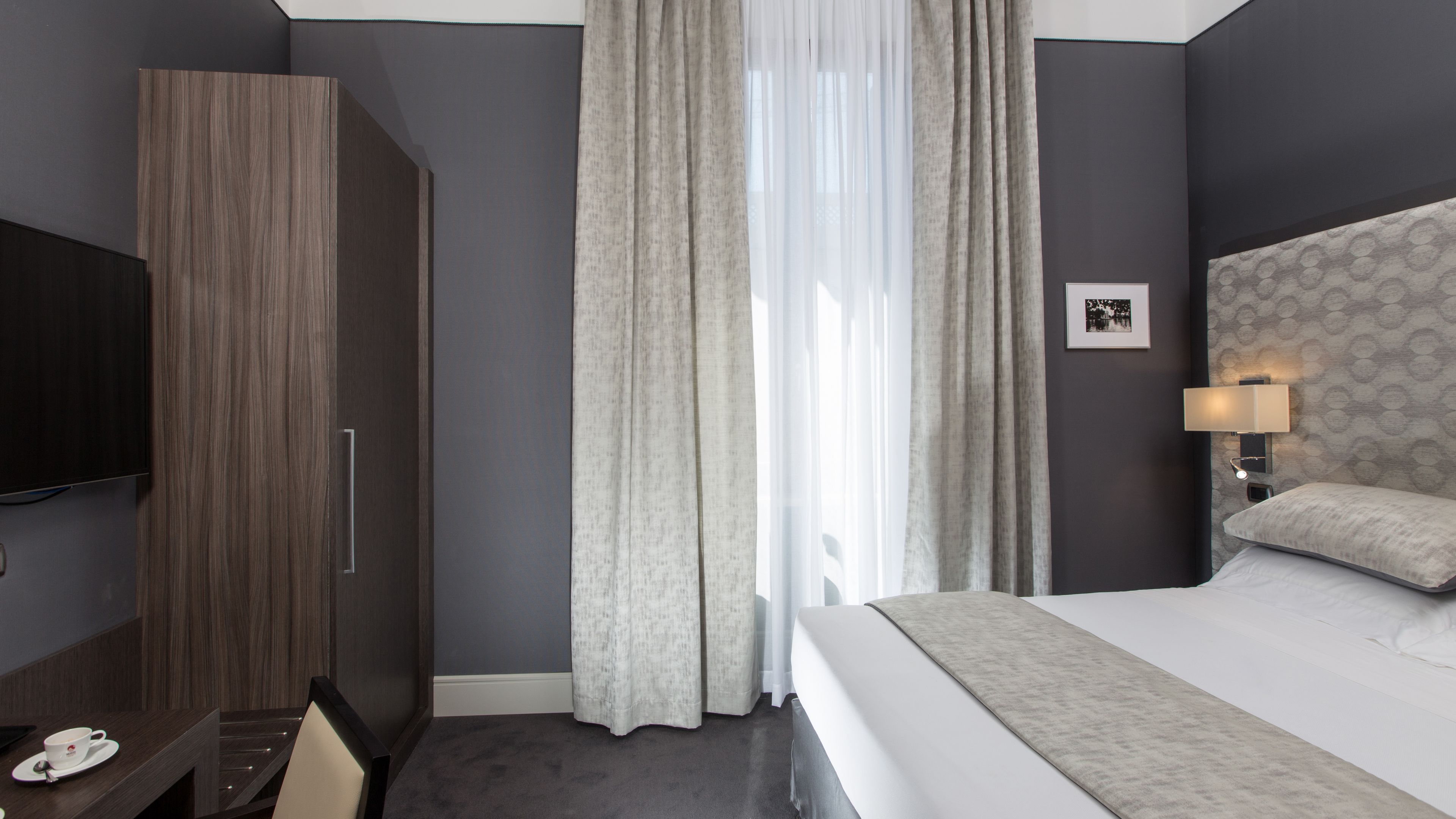A modern hotel room with a large bed, a TV, a desk with a cup, and a window with curtains.