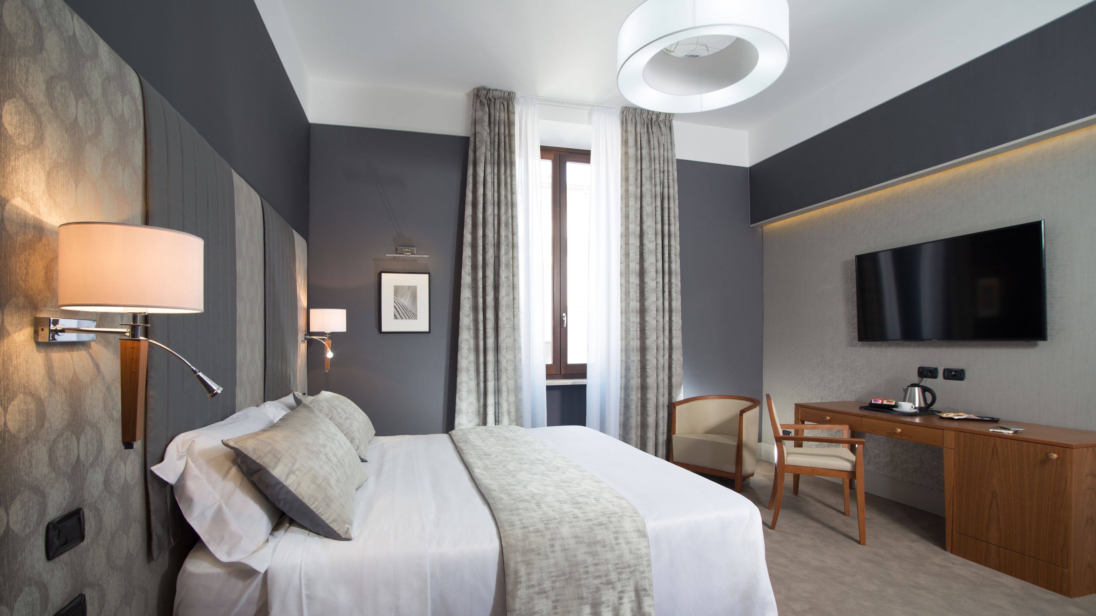 A modern hotel room with a large bed, a desk, a chair, a flat-screen TV, and a window with curtains.
