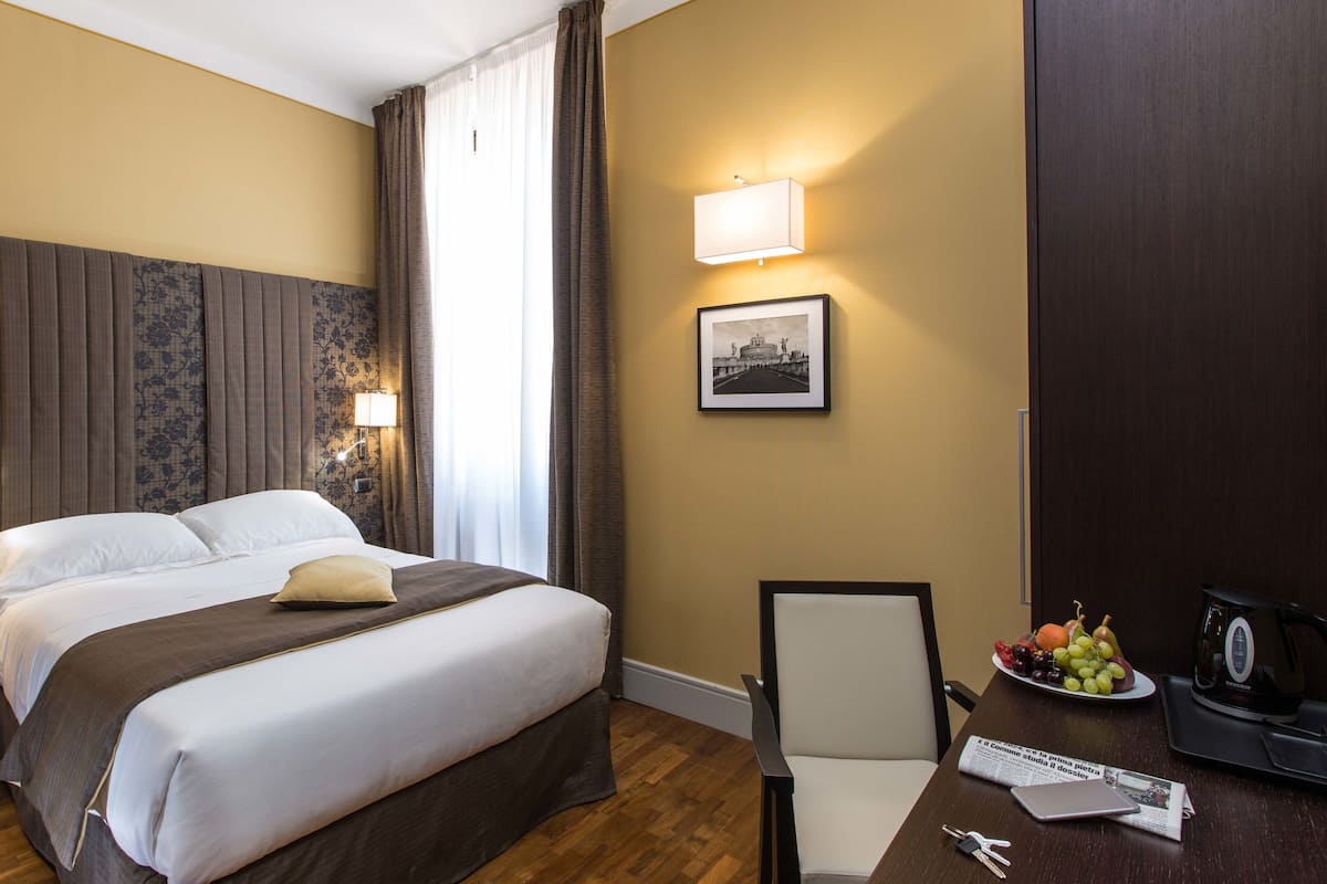 Une chambre d’hôtel avec un grand lit, deux tables de chevet, un bureau avec une chaise, une lampe, un tableau encadré et une assiette de fruits.