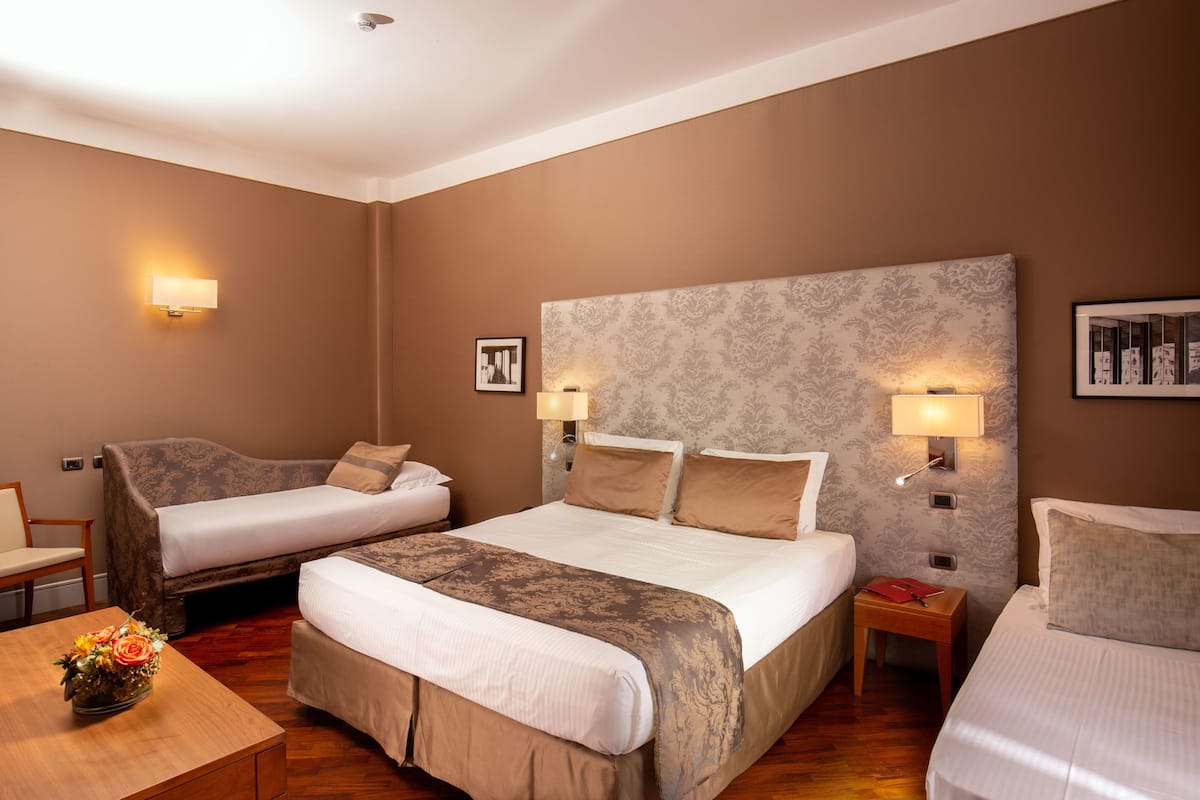 Une chambre d’hôtel avec deux lits, un canapé, une petite table avec un vase et des lampes fixées au mur.