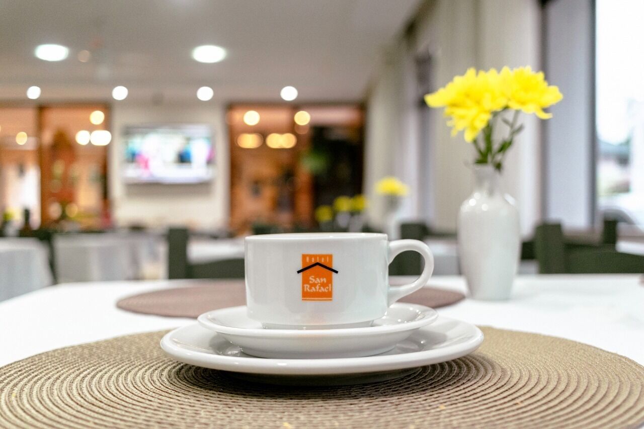 A hotel room with a dining table set for two, a white cup with a logo, and a vase with yellow flowers.