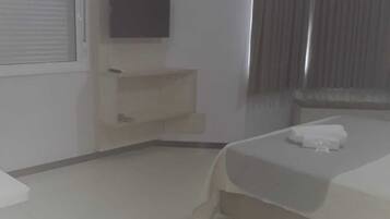 Habitación de hotel con una cama, un televisor de pantalla plana montado en la pared, una mesita de noche pequeña y una ventana con cortinas.