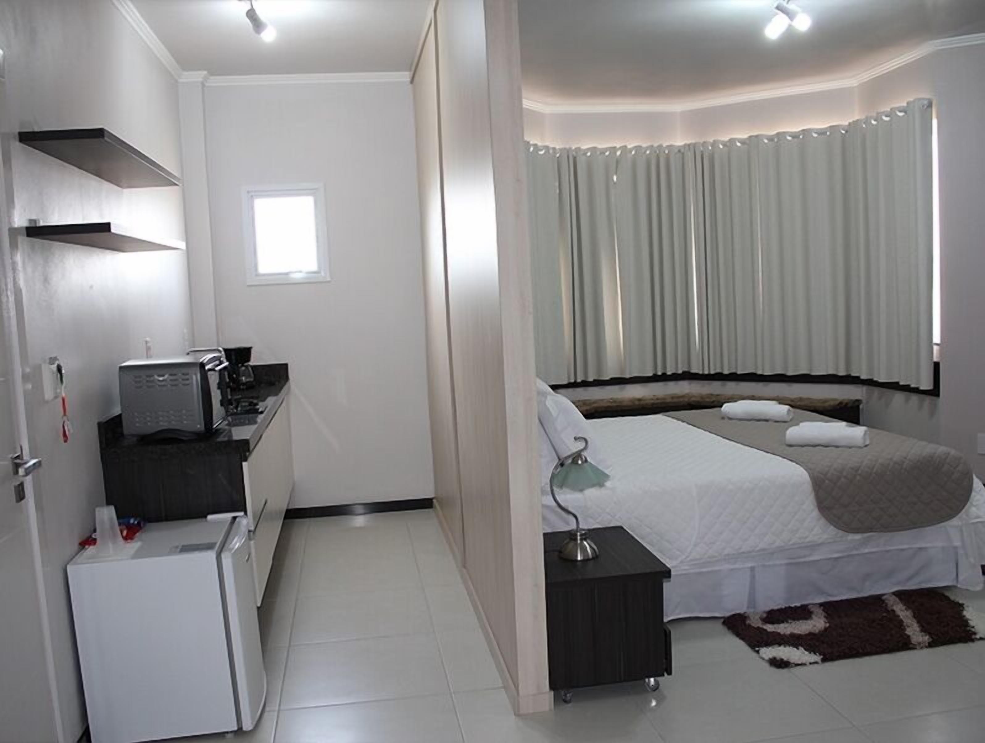 A compact hotel room with a kitchenette, a bed with a bedside table, and a window with curtains.