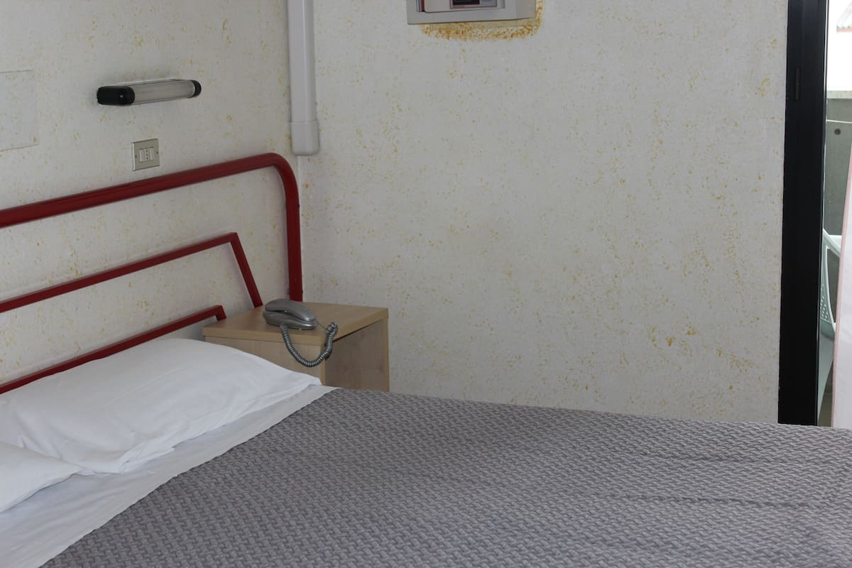 A hotel room with a single bed, a bedside table with a telephone, and a wall-mounted light switch.