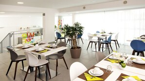 Een moderne eetruimte met witte tafels, blauwe en grijze stoelen en een plant in een pot in het midden.