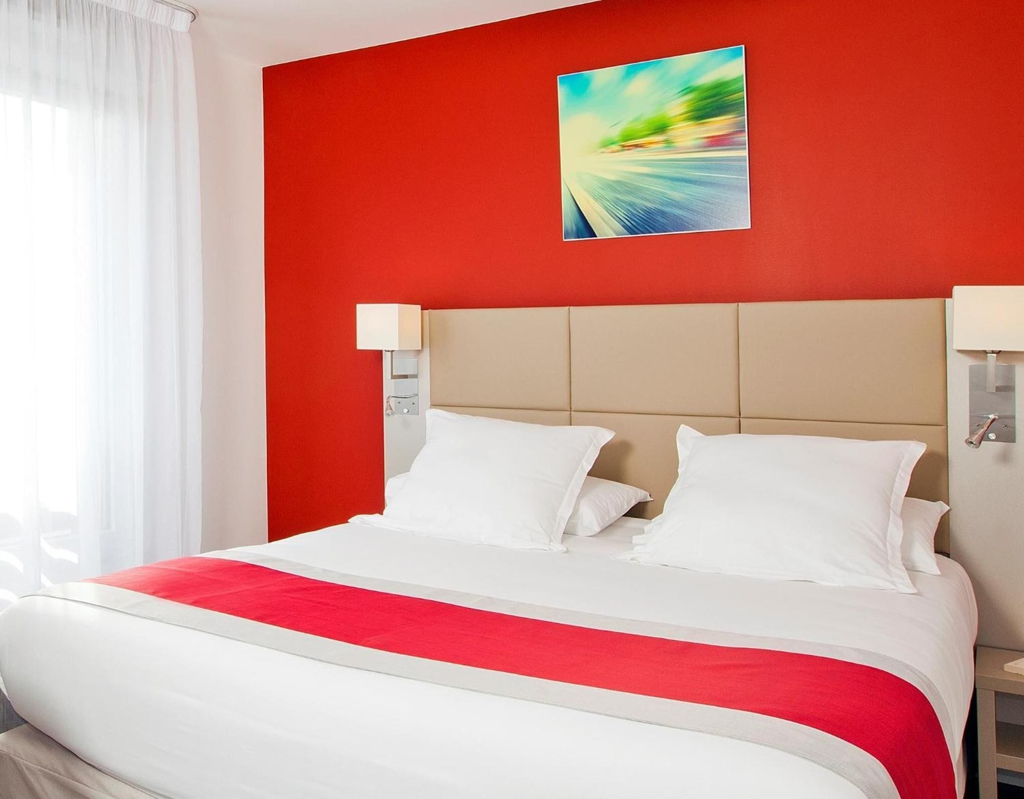 A hotel room with a red accent wall, a large bed with white bedding, and a modern abstract painting above the bed.