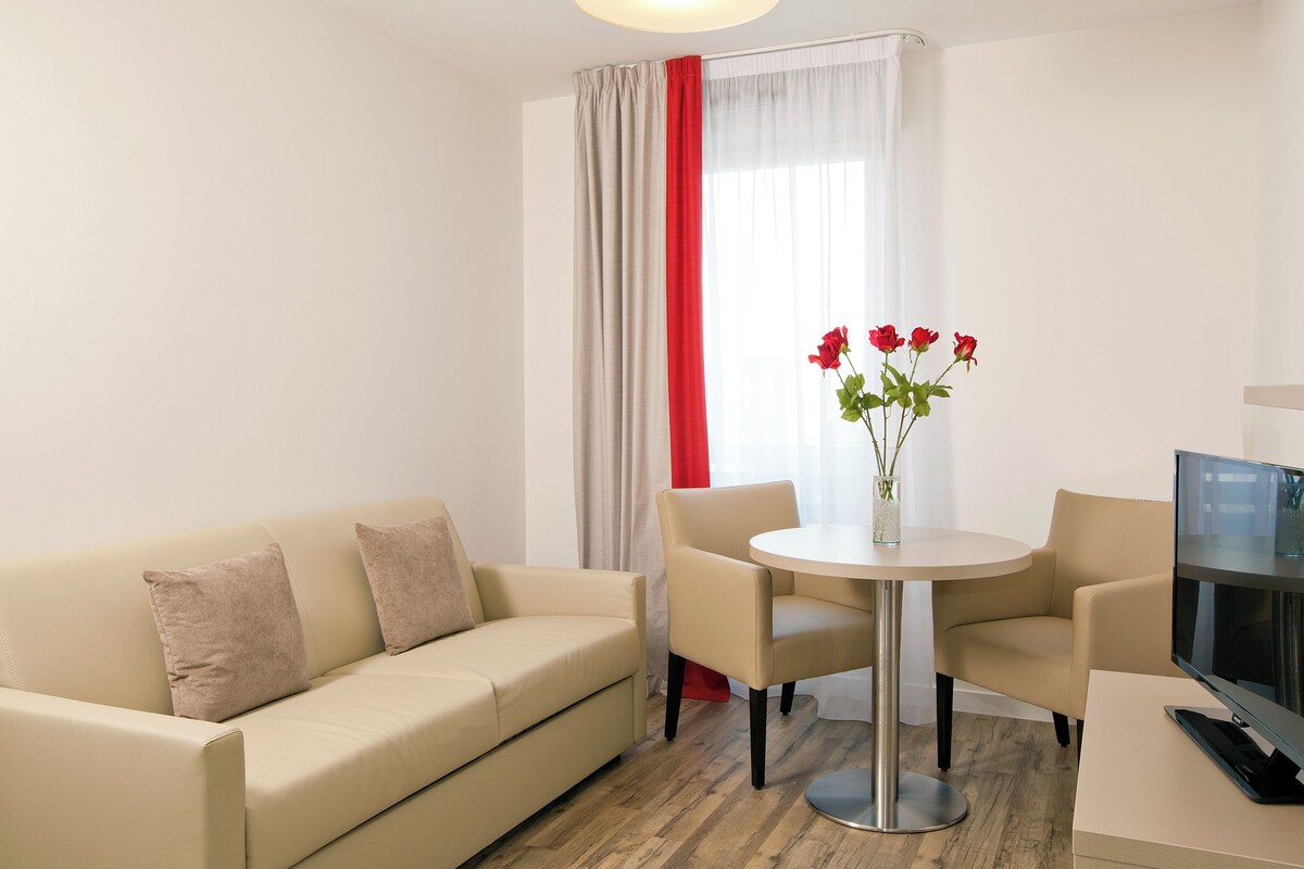 Una sala moderna con un sofá color beige, dos sillones, una mesa redonda y un televisor sobre un mueble.