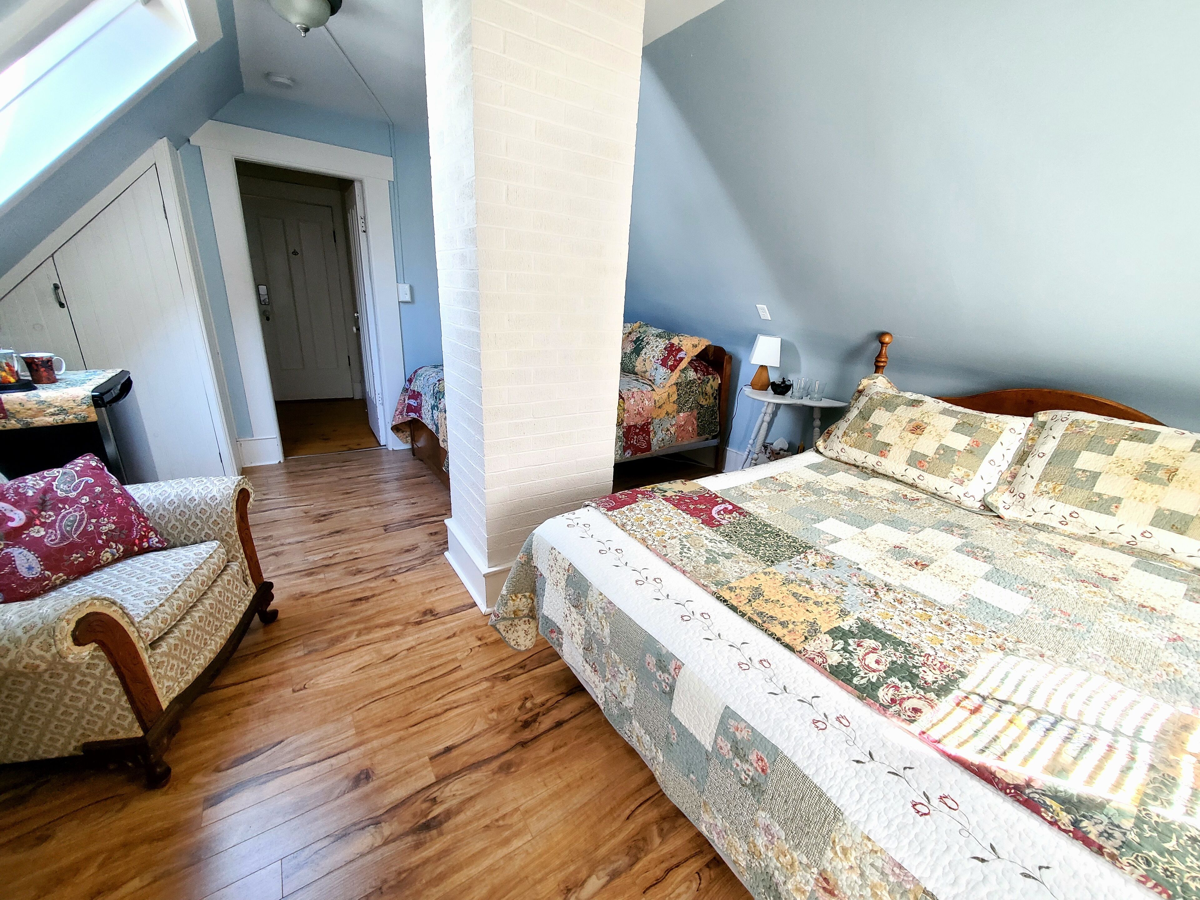 A bedroom with a wooden floor, a bed with a floral quilt, a chair with a red cushion, and a small table with a lamp.