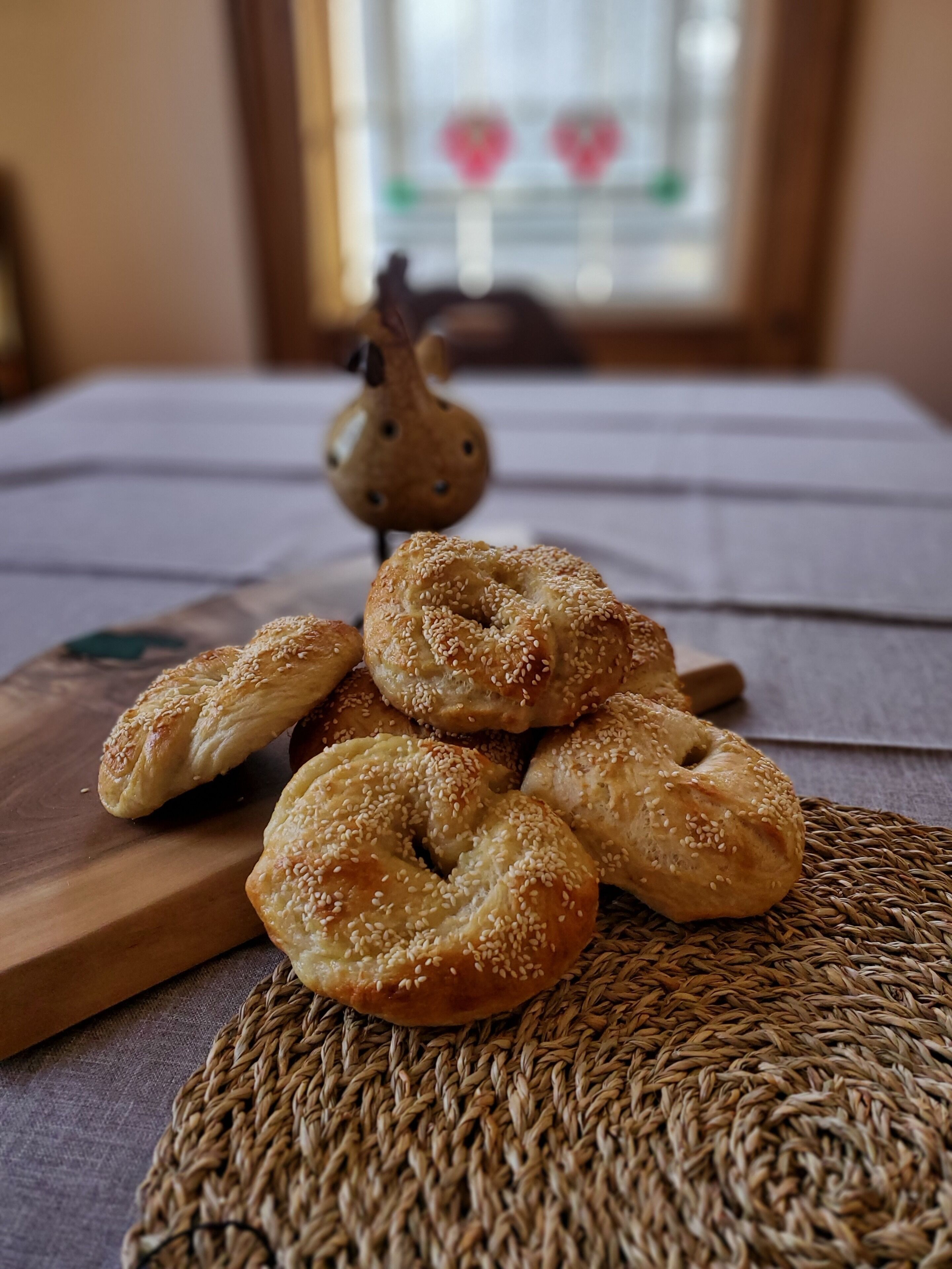 A table with several sesame seed bagels on a woven placemat, with a wooden cutting board and a decorative bird figurine in the background.