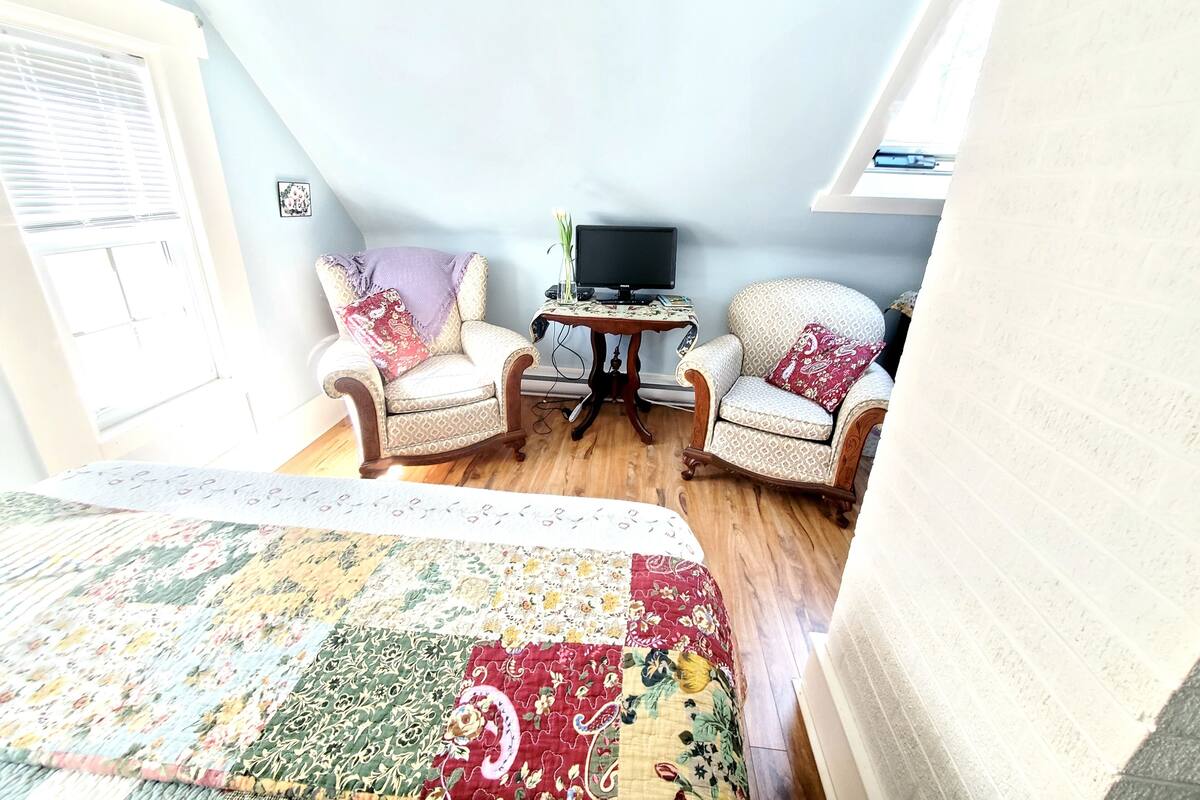 A cozy room with a large bed, two patterned armchairs, a small table with a TV, and a window with blinds.