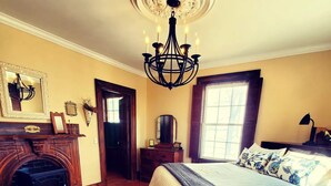 A bedroom with a large bed, a fireplace, a chandelier, and a window with curtains. - Fisherman's Daughter Bed & Breakfast (Mahone Bay)