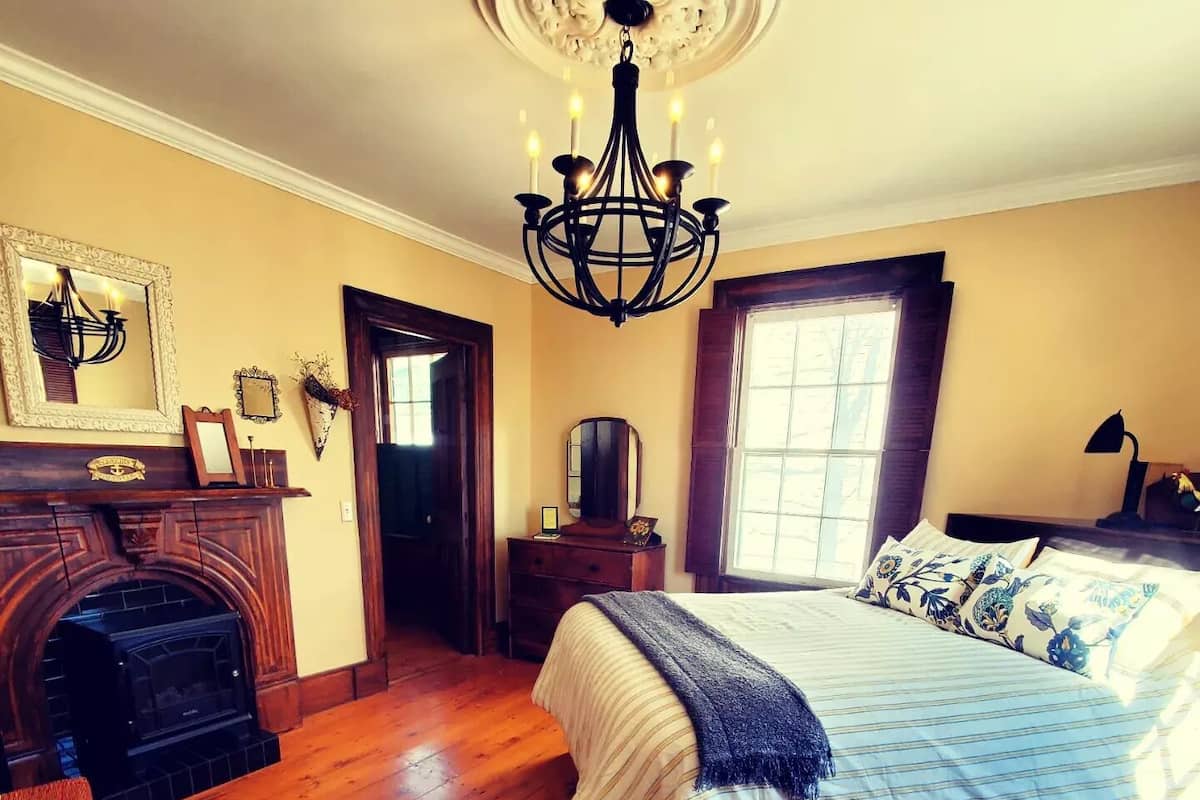 A bedroom with a large bed, a fireplace, a chandelier, and a window with curtains.