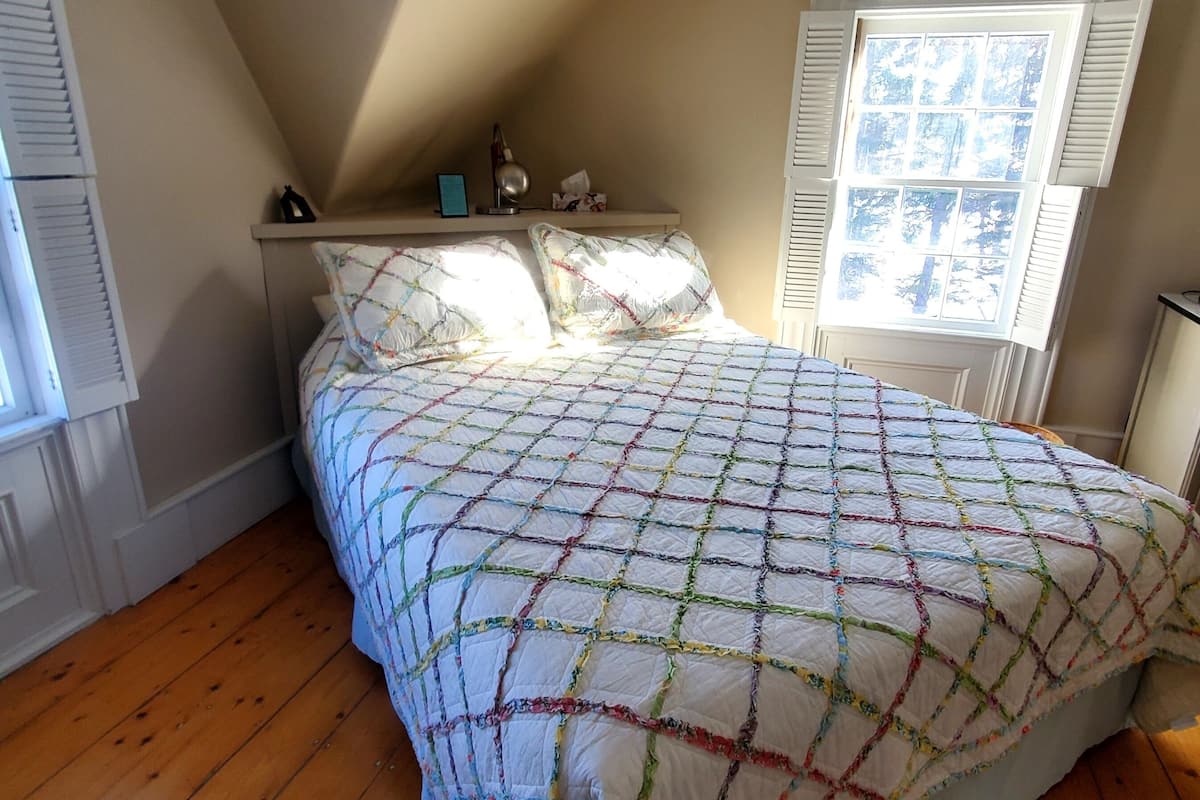 A bedroom with a sloped ceiling, wooden flooring, a bed with a colorful quilt, a nightstand, a window with shutters, and a ceiling fan.