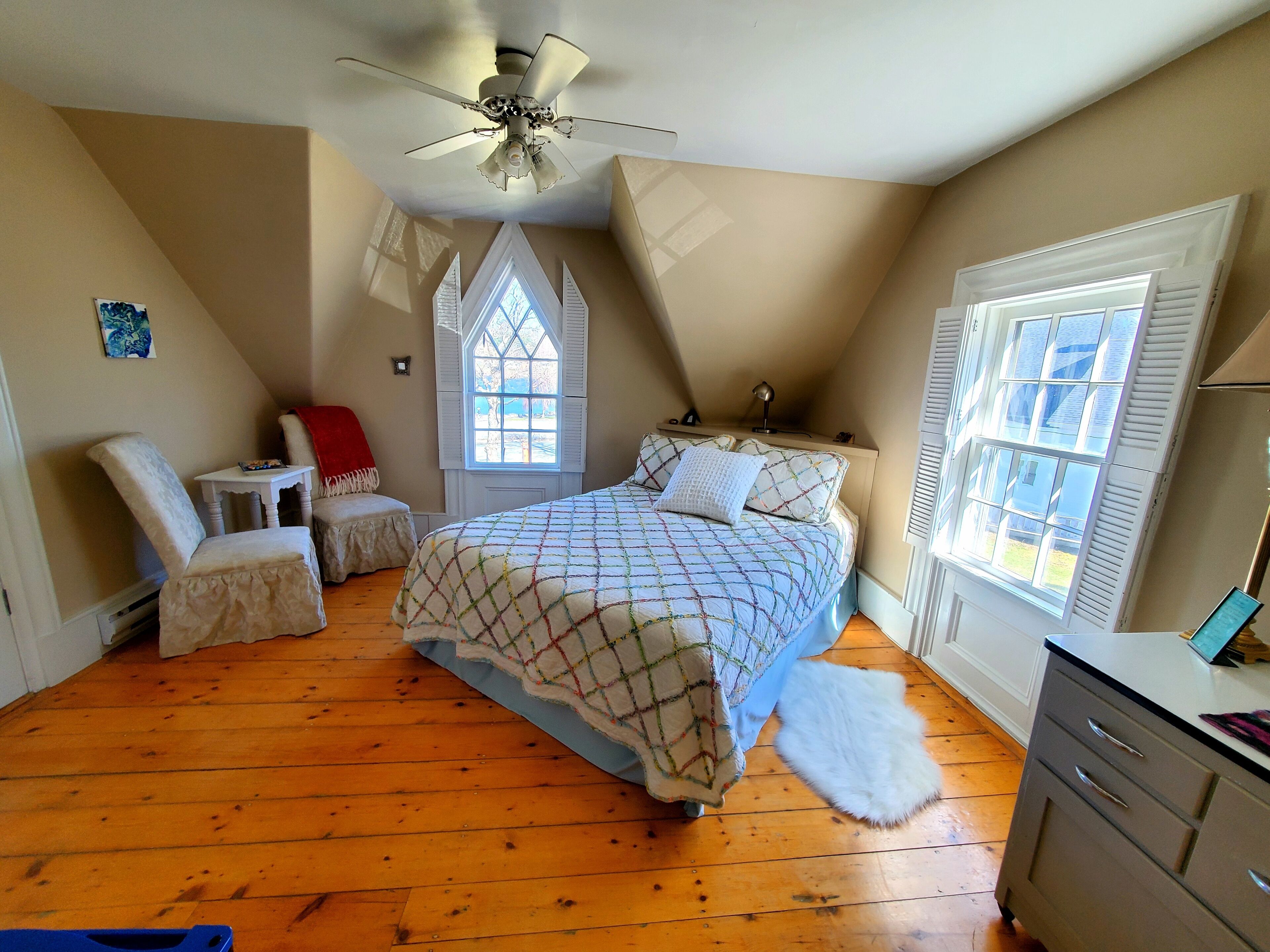 A bedroom with a bed, a chair, a small table, a heater, a window, and a ceiling fan.