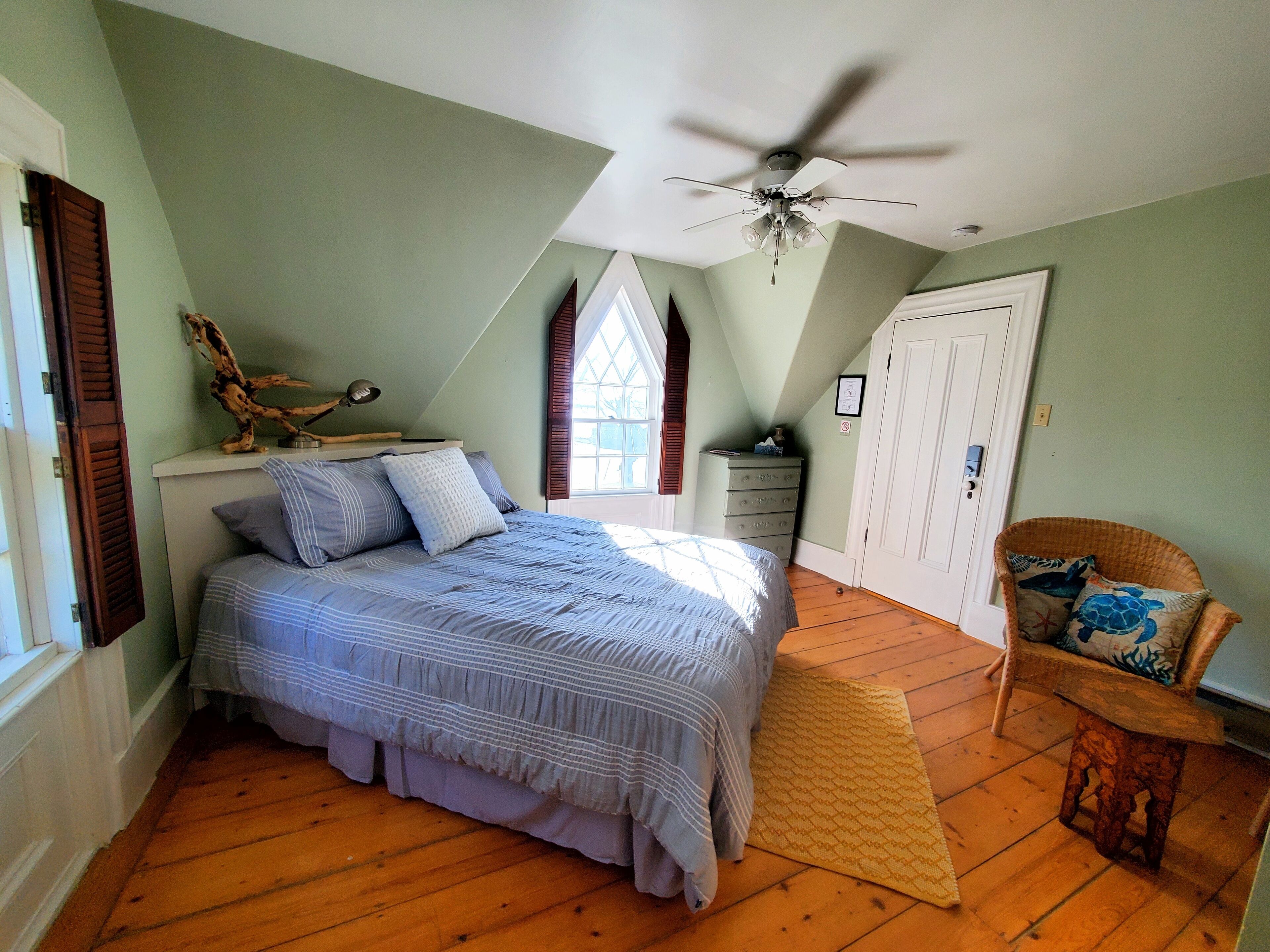 A bedroom with a bed, a ceiling fan, a dresser, a chair, and a window with shutters.