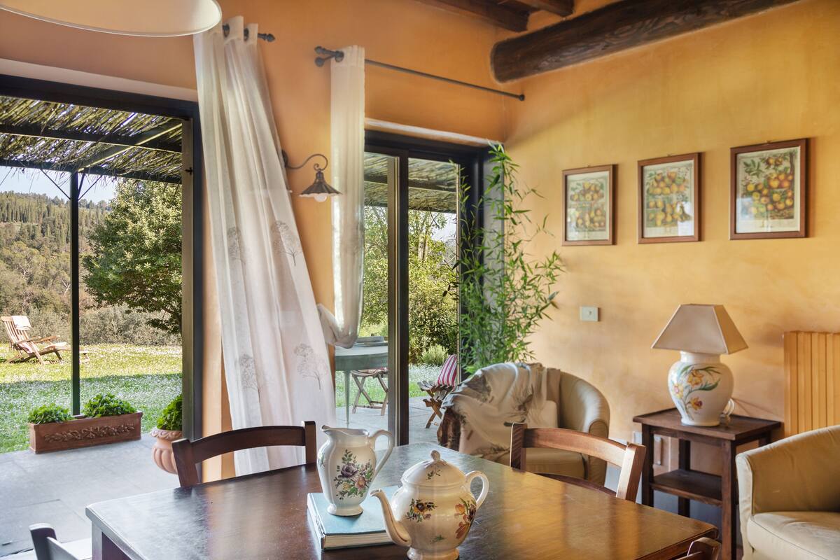 A dining area with a table set for two, a pitcher, a teapot, and a lamp on a side table. There is a view of a garden through a glass door.