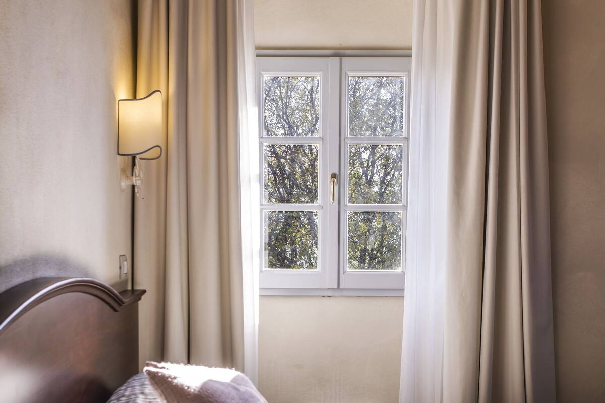 A neatly made bed with striped bedding and a single pillow, a bedside lamp, and a window with curtains.