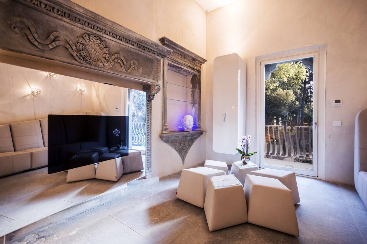 A modern living room with a flat-screen TV, a fireplace with a blue light, and a balcony with a view of trees.