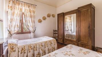 A bedroom with a bed, a wooden wardrobe, a window with curtains, and a small bedside table.
