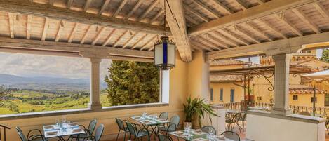 An outdoor dining area with tables and chairs, a view of the countryside, and a rustic wooden ceiling.
