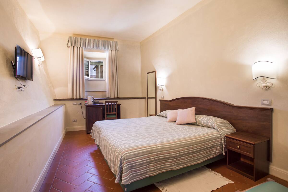 A bedroom with a bed, bedside table, nightstand, TV, and a window with curtains.