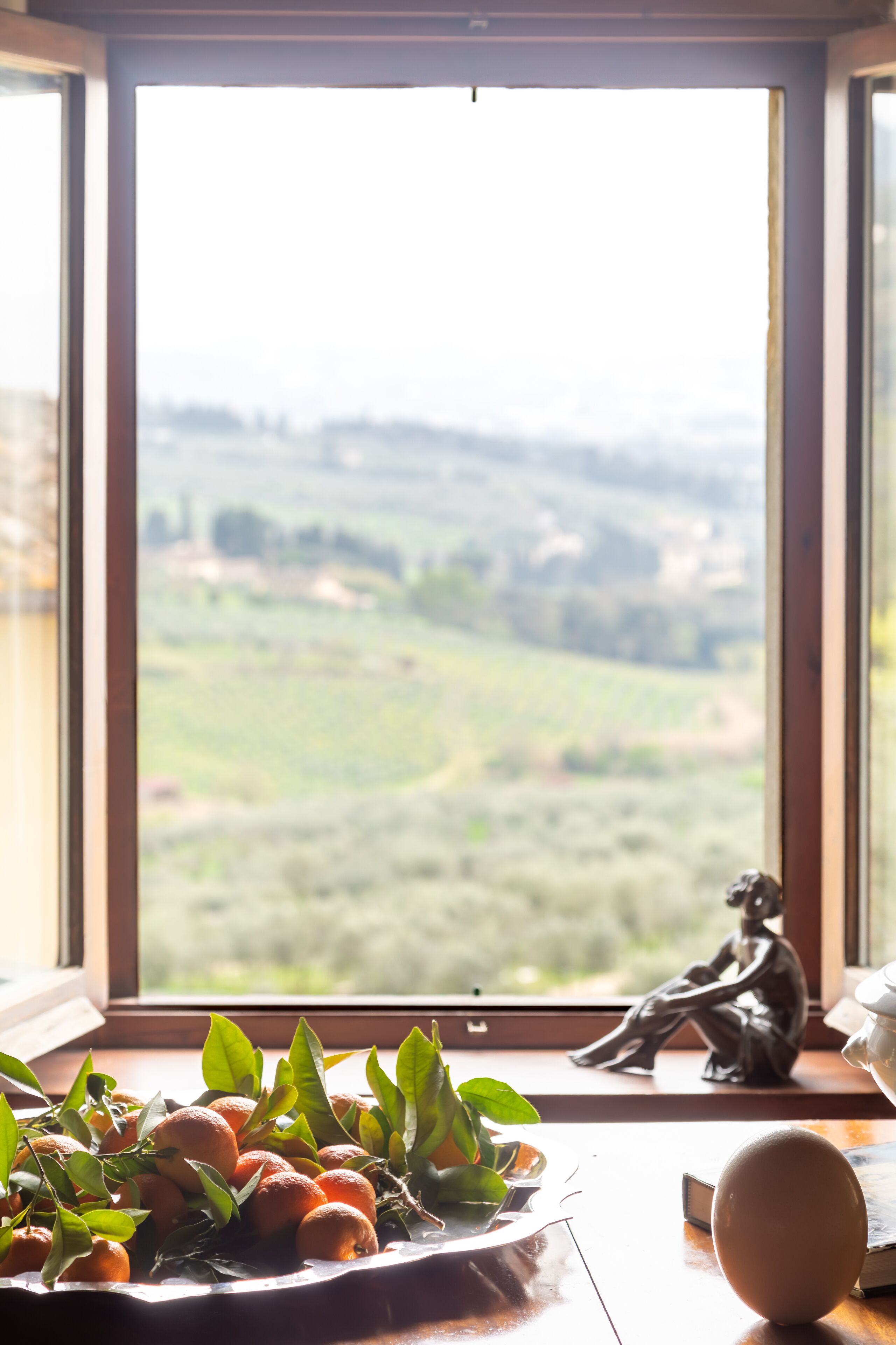 A table with oranges and a ceramic sculpture by a window with a view of a green landscape.