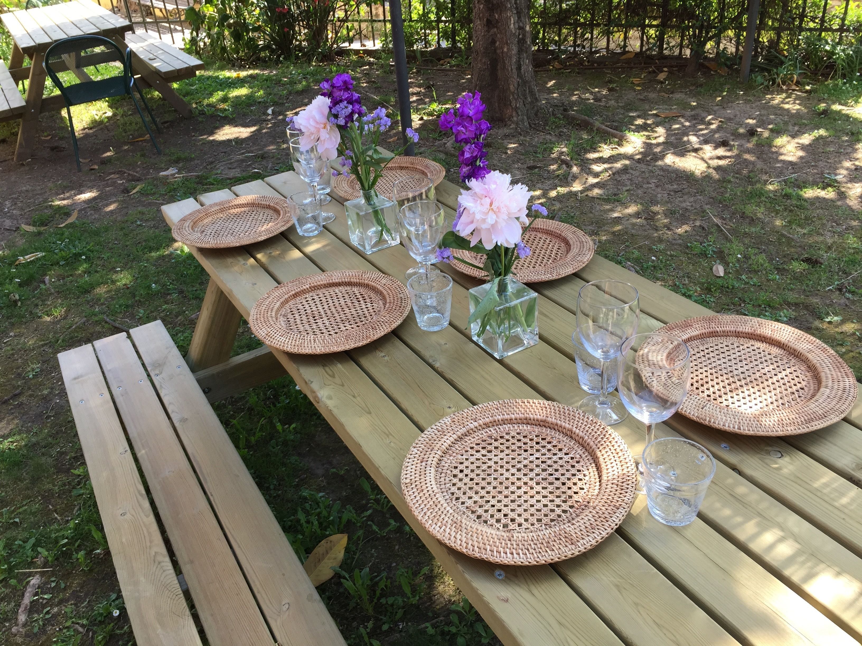 Un juego de mesa de picnic de madera con individuales tejidos, vasos transparentes y pequeños jarrones con flores.