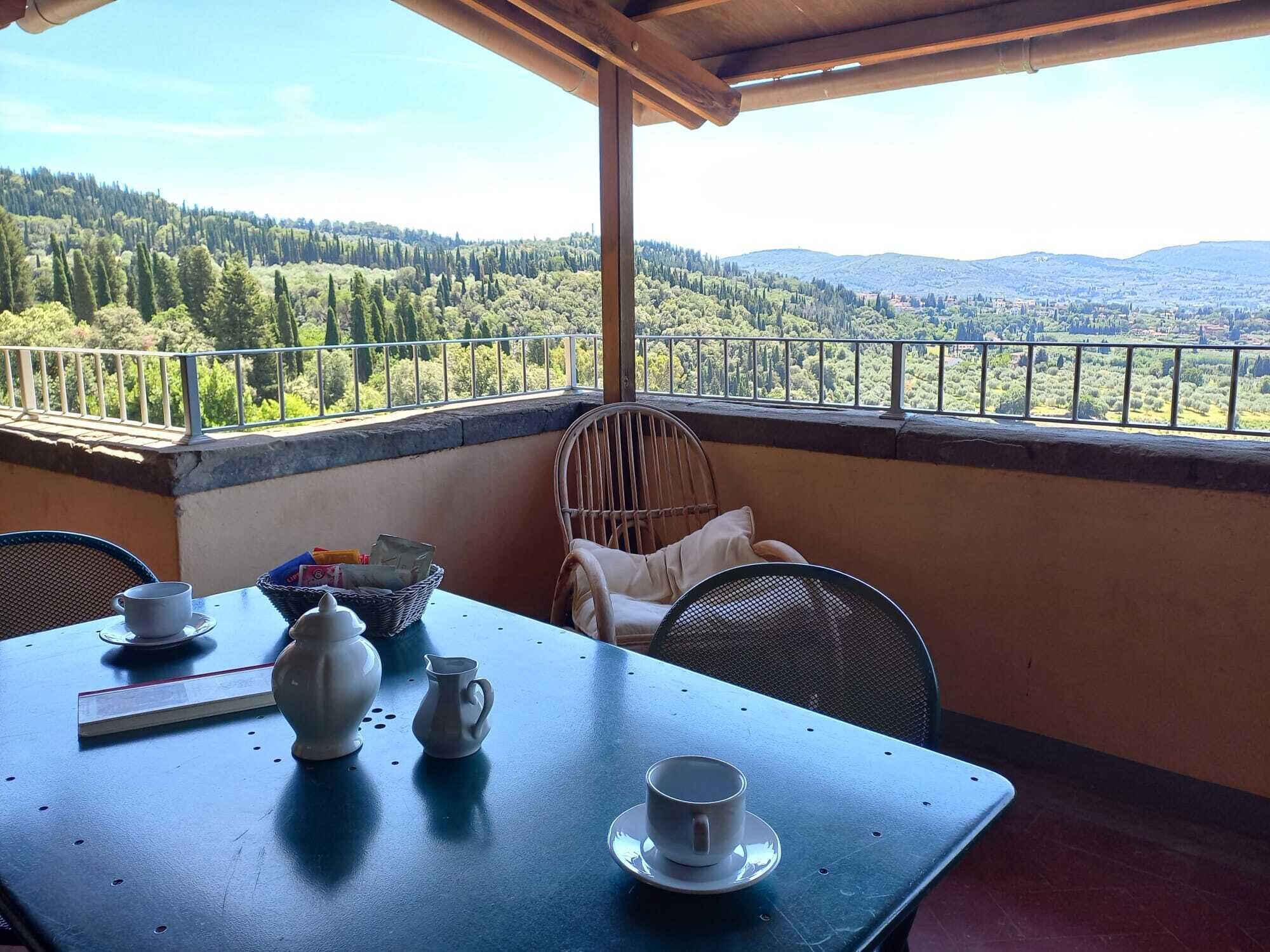 Un balcone con un tavolo apparecchiato per due persone, una sedia in vimini e vista su un paesaggio boschivo.