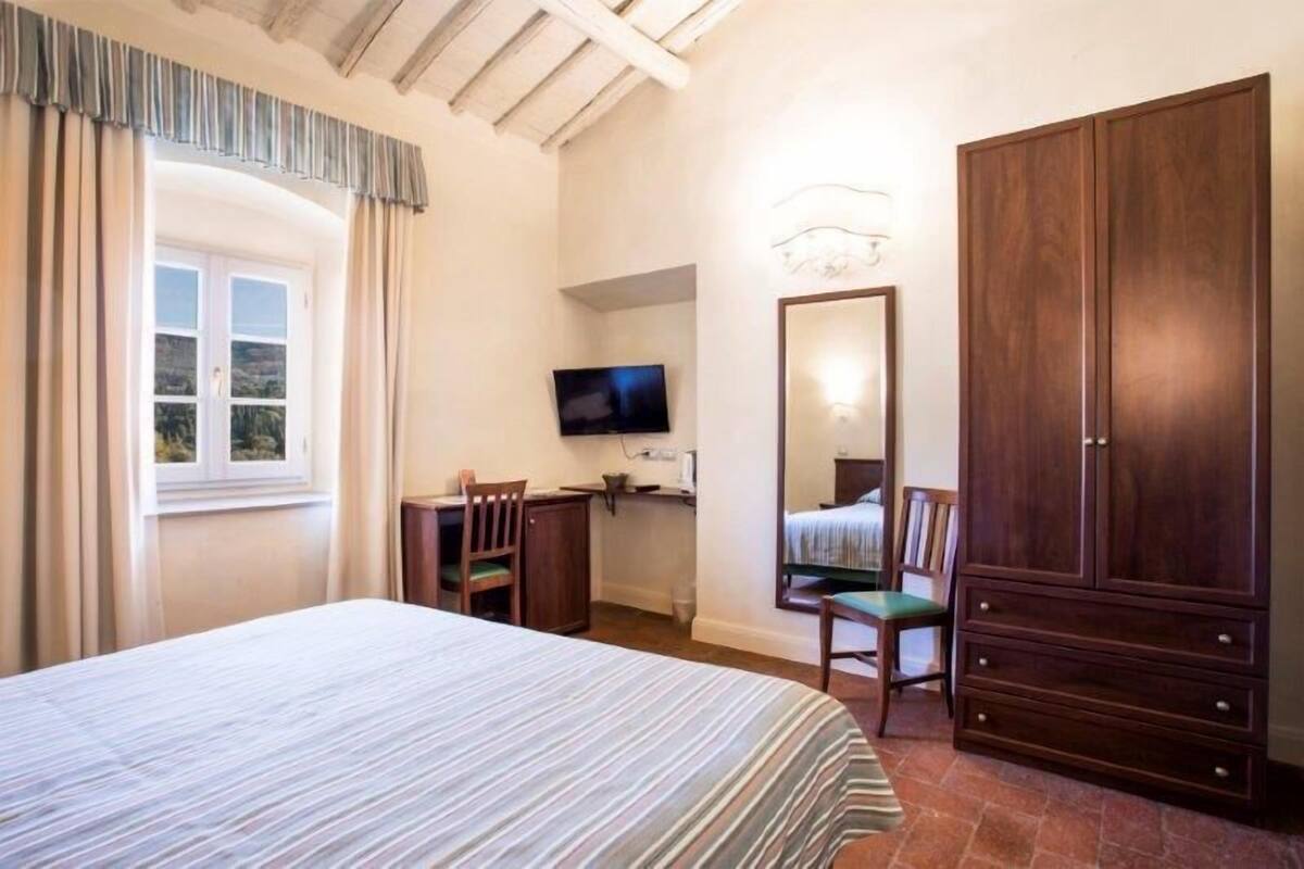 A bedroom with a bed, a desk, a chair, a television, a wardrobe, and a window with curtains.