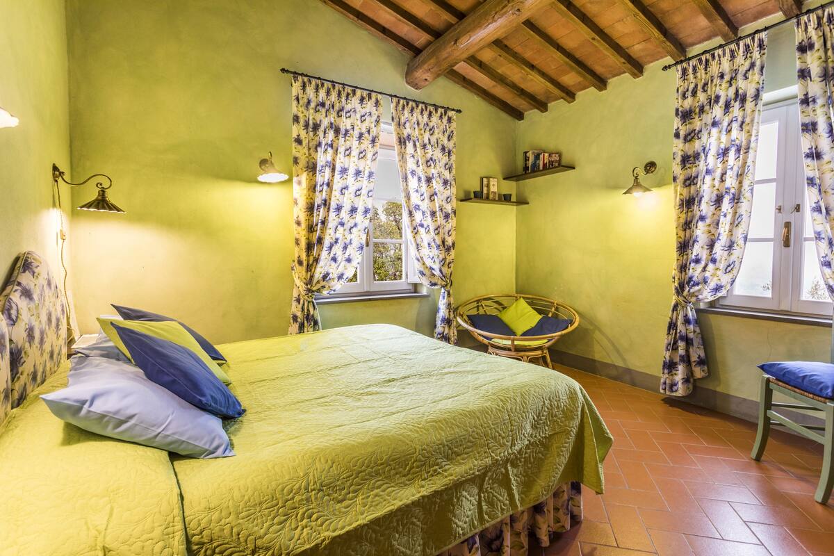 A room with a bed, a small table, and a chair. There are curtains on the windows, and a shelf with books.