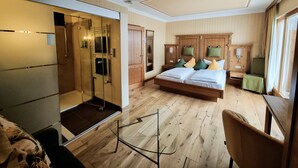 Habitación de hotel con piso de madera, una cama con muchos almohadones, armario de madera, cabina de ducha de vidrio y una pequeña mesa con una silla.