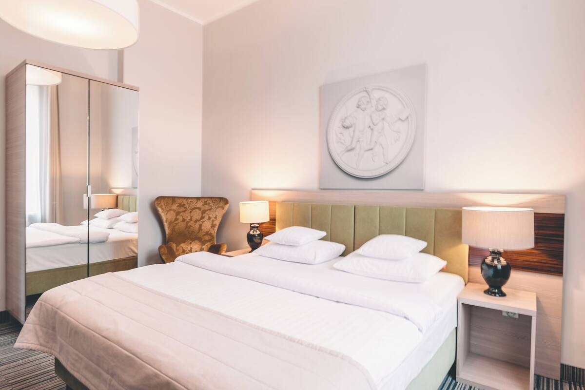 A hotel room with a large bed, two bedside tables with lamps, a chair, a mirror, and a wall-mounted artwork.