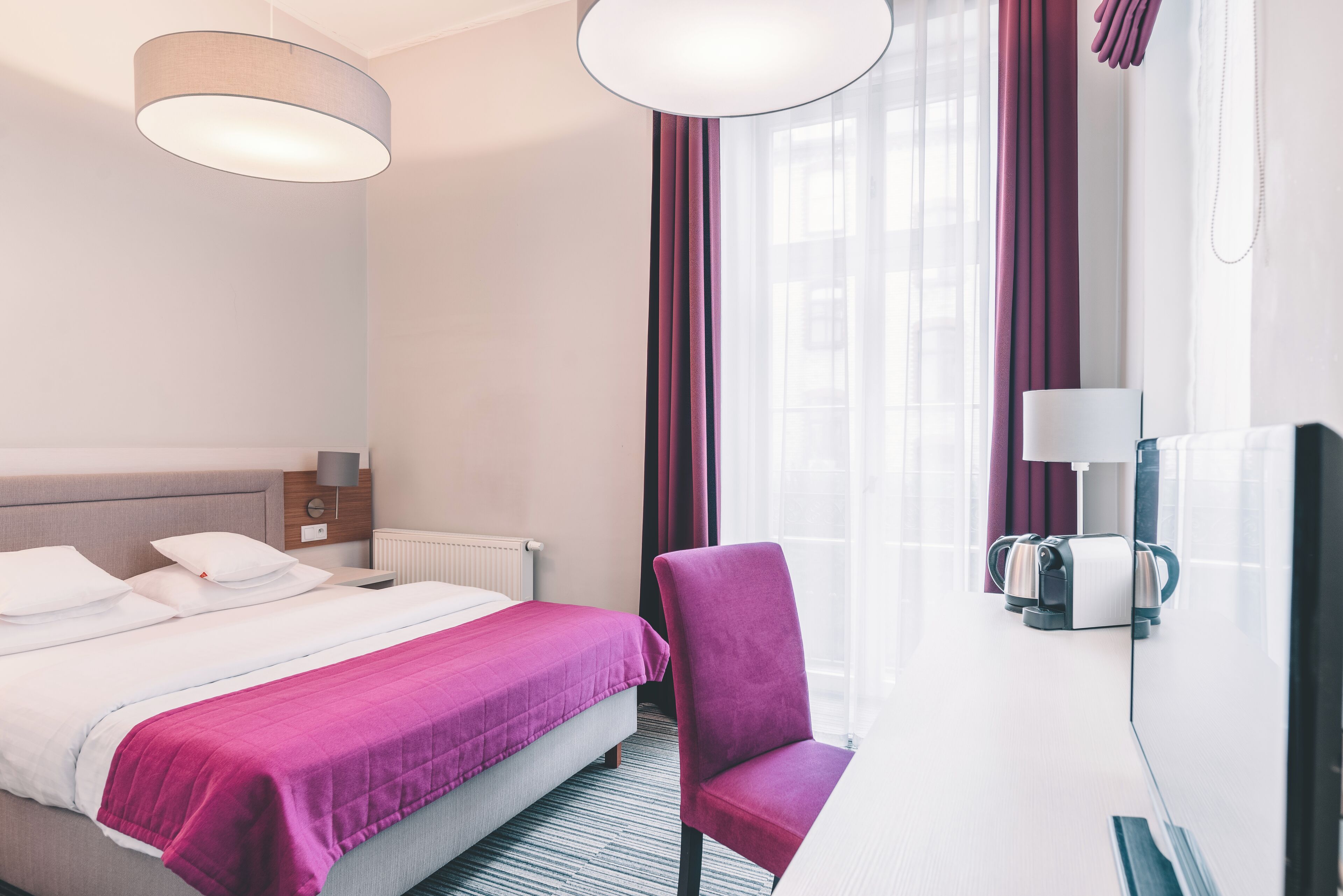 A hotel room with a bed, a chair, a desk with a lamp, a mirror, and a window with curtains.